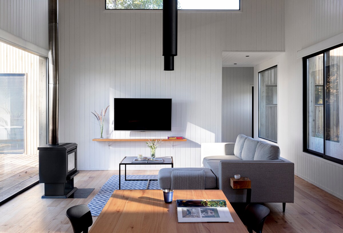 A bright and spacious living area features large windows allowing natural light to illuminate the room. A minimalist gray sofa is positioned alongside a wooden table, while a black wood stove adds warmth. A television is mounted on the wall, creating a modern ambiance.