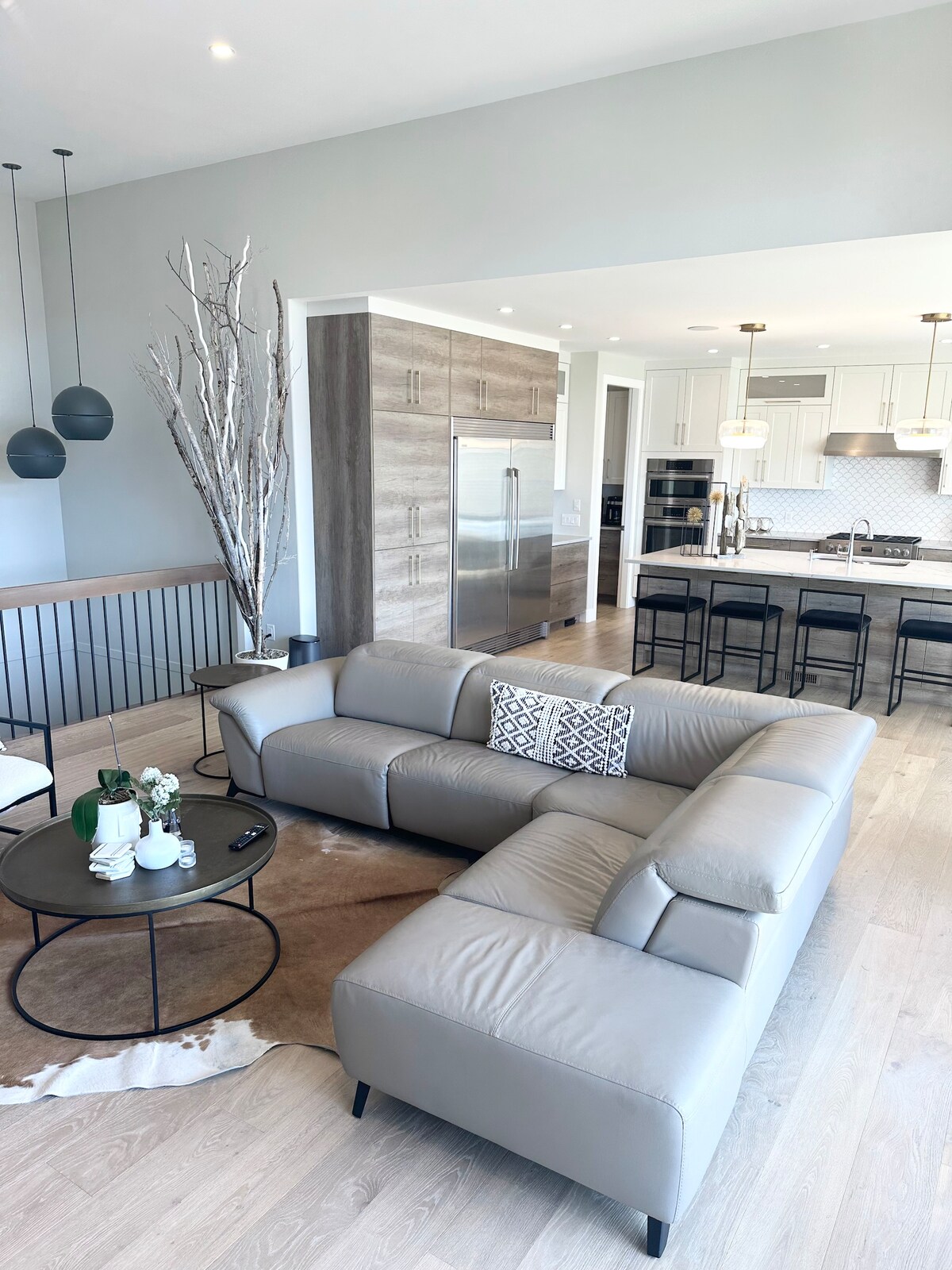 A modern living area is presented with a light grey sectional sofa, centered on a round coffee table with decorative items. The kitchen is visible in the background, featuring sleek cabinetry and stainless steel appliances. Pendant lights hang from the ceiling, enhancing the open layout.