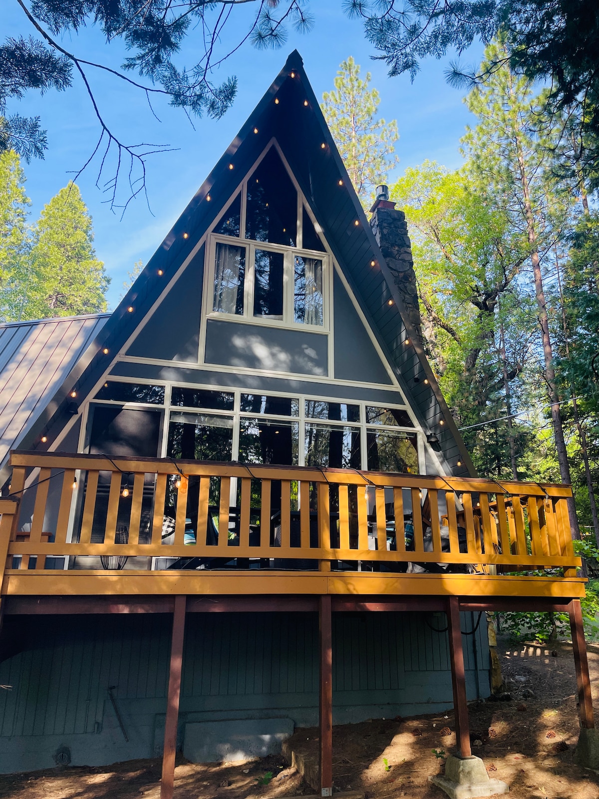 A cozy A-frame cabin stands among the trees, showcasing large windows that invite natural light into the living space. A wraparound deck features a yellow railing, providing outdoor space for seating. String lights are visible, enhancing the cabin's charm during twilight hours.