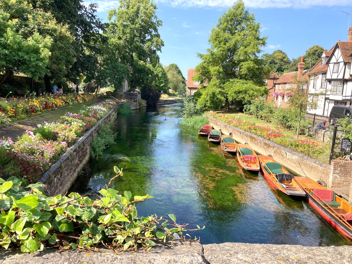 The scenic view includes a calm river bordered by lush greenery and vibrant flower beds. Rowboats can be seen lined along the water's edge, creating a tranquil atmosphere. Historic buildings with charming wooden façades are positioned in the background, blending with the natural surroundings.