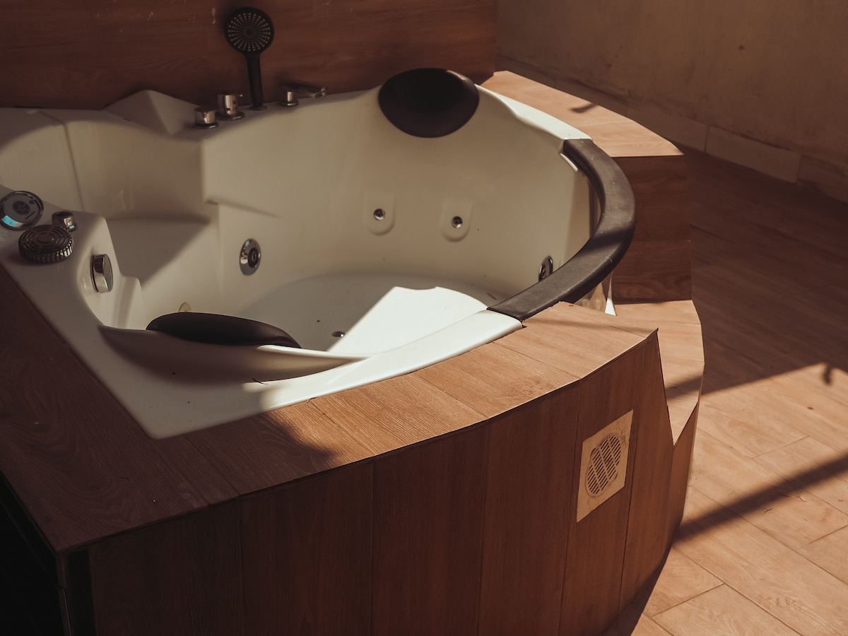A modern whirlpool tub is positioned within a minimalist setting, featuring smooth surfaces and a warm wood finish. Sunlight casts gentle shadows, highlighting the sleek curves and inviting design of the tub.