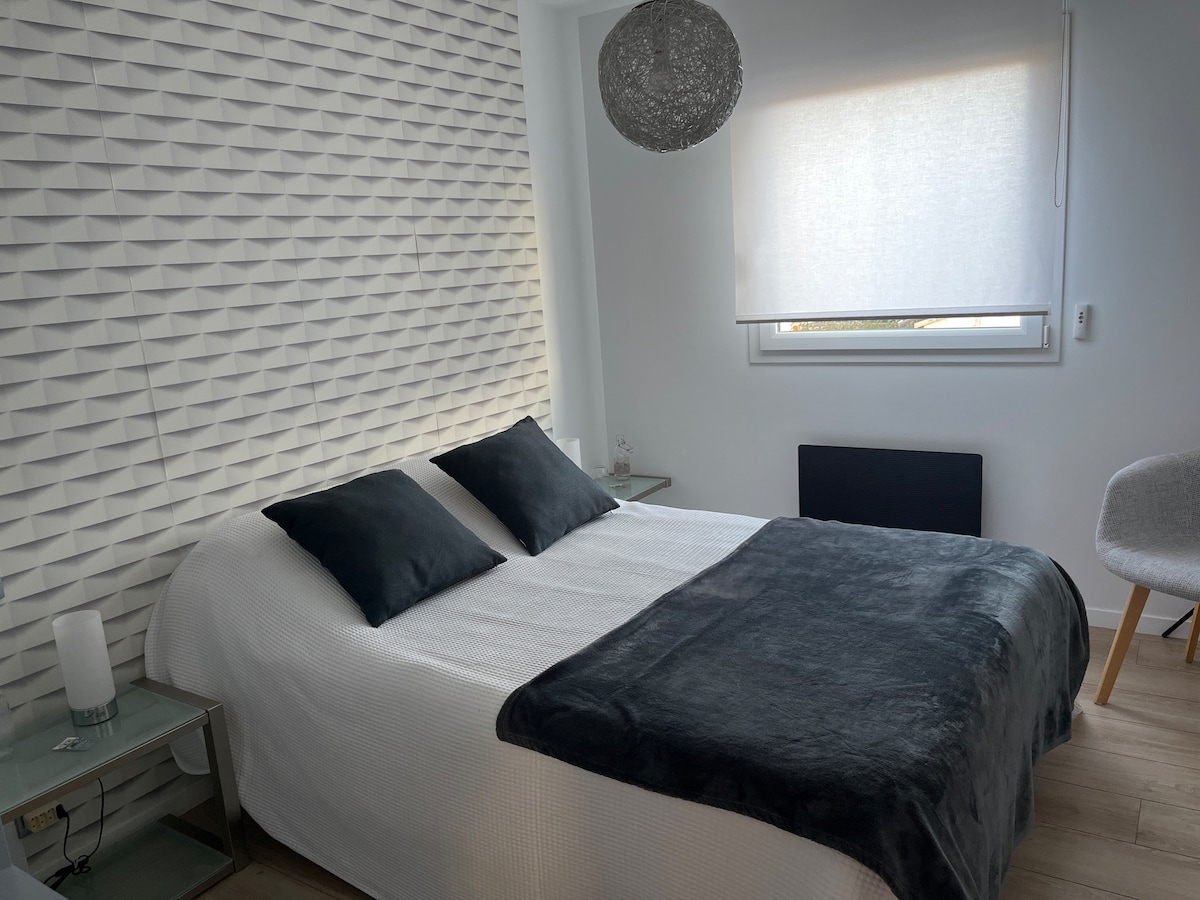 A cozy bedroom is presented with a textured white feature wall. A spacious bed is adorned with dark cushions and a soft blanket. A small bedside table is positioned on each side, while a modern lamp and a stylish chair enhance the serene atmosphere.
