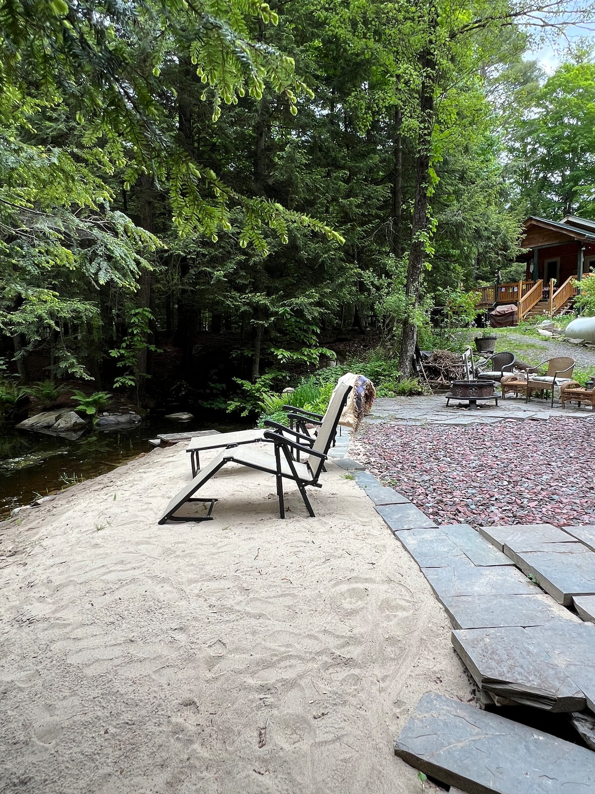 A sandy area is visible next to a small pond, featuring black outdoor chairs. Surrounding greenery includes tall trees, creating a natural setting. A stone path leads to a fire pit with additional seating, suggesting a welcoming space for relaxation.