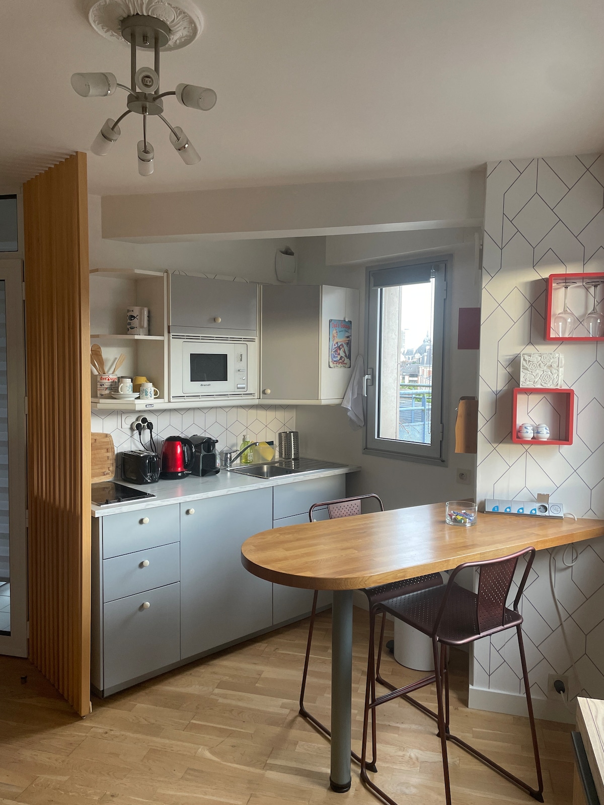 A compact kitchen area is showcased, featuring modern appliances including a microwave and kettle. A round wooden table with metal legs is accompanied by two chairs. Natural light is let in through a window, illuminating the space with a warm ambiance.