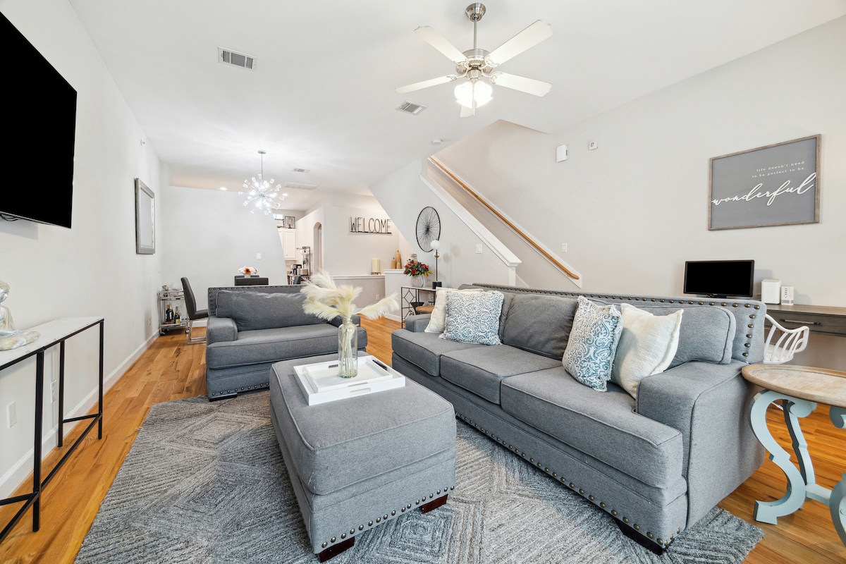 A spacious living area is furnished with a large gray sectional sofa, accented by decorative pillows. A coffee table rests in the center, while a flat-screen TV is mounted on the wall. Light wooden floors contribute to the bright and airy atmosphere.