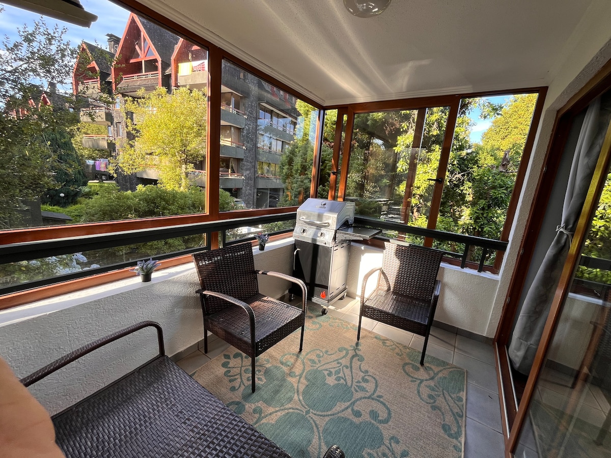 A screened terrace features two wicker chairs positioned around a small table. A gas grill is situated nearby against a backdrop of greenery and residential buildings visible through large windows. The space is adorned with a patterned rug that adds a touch of comfort.