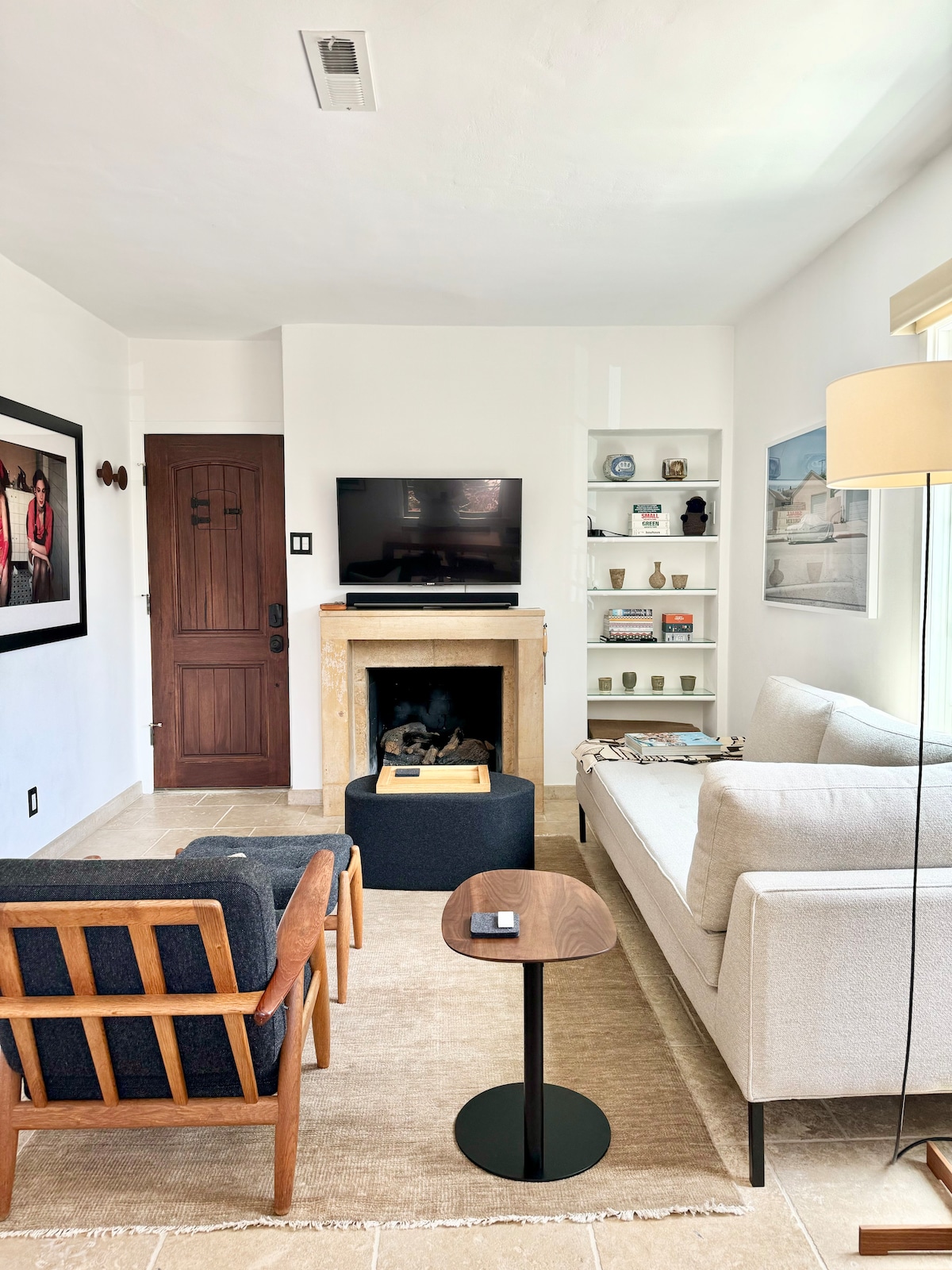 The living area features mid-century modern furnishings, including a light-colored sofa and accent chairs. A gas fireplace is surrounded by a simple mantel, and a smart TV is mounted above it. Decorative shelves display local ceramics and art, enhancing the inviting atmosphere.