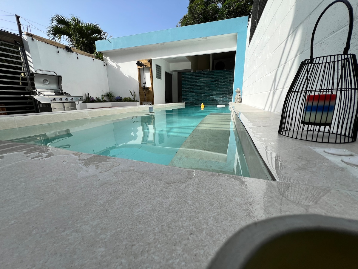 A refreshing swimming pool is framed by smooth stone decking, with a modern outdoor area visible in the background. Clear water reflects the sky, and a hint of colorful decor is present near the poolside, adding a subtle touch to the serene setting.