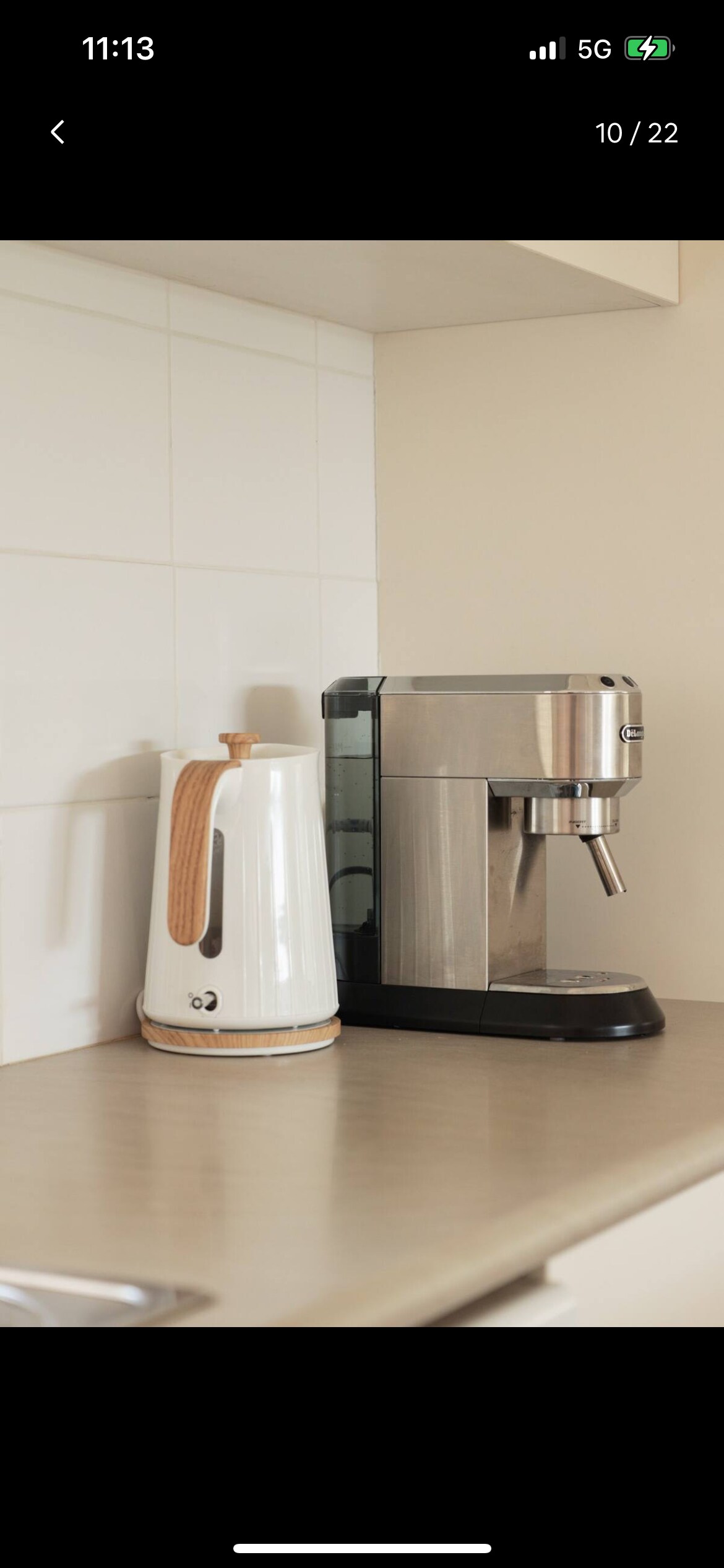 A sleek kitchen countertop is showcased, featuring a modern stainless steel coffee machine beside a white kettle with a wooden handle. The neutral-colored tiles create a clean and functional atmosphere, emphasizing the appliances' contemporary designs.