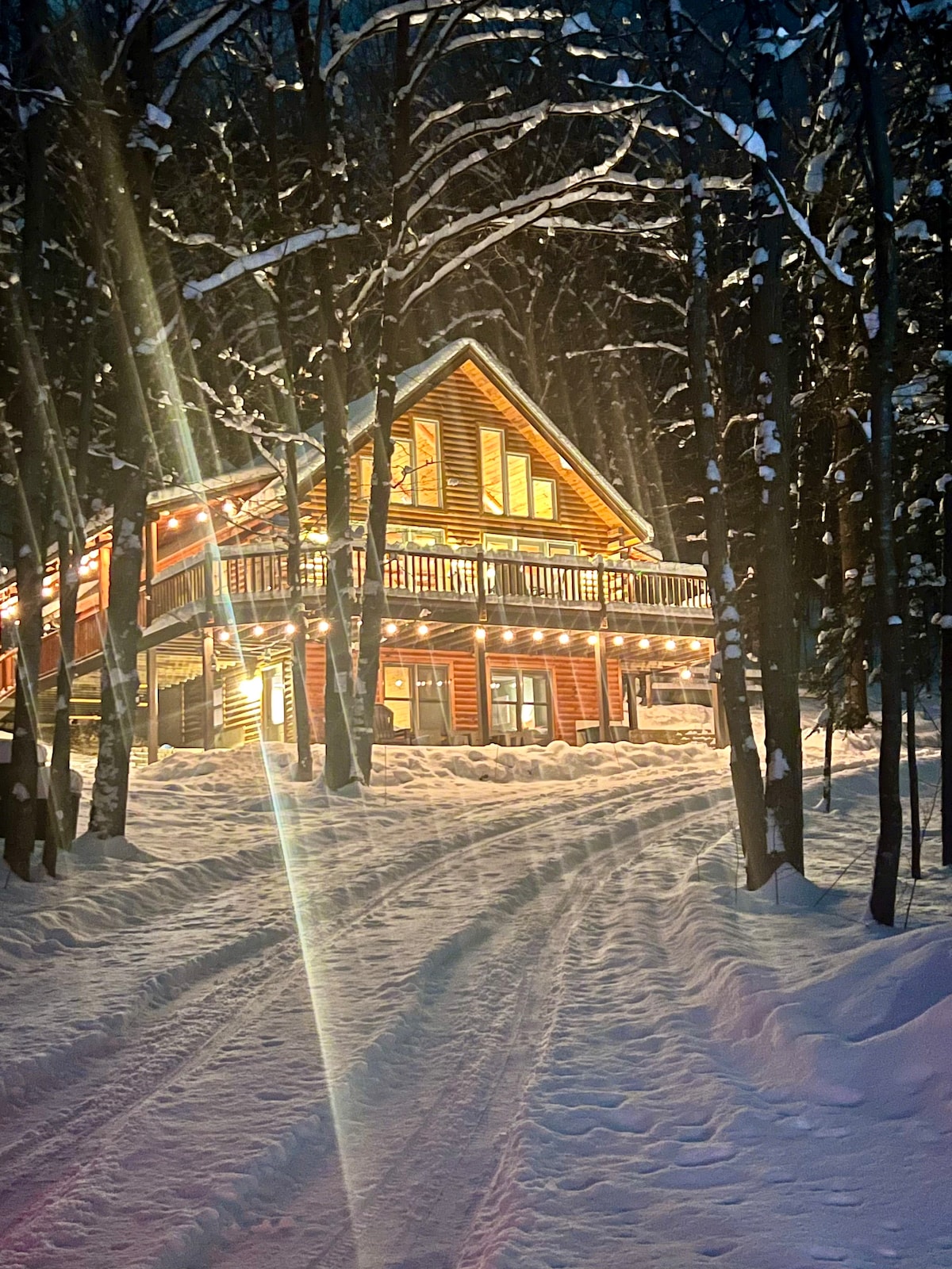 A log cabin is nestled in a snowy landscape, illuminated by warm lights. Tall trees frame the path leading to the cabin, while snow softly blankets the ground. The inviting exterior features a wrap-around deck and numerous windows that reflect the evening glow.