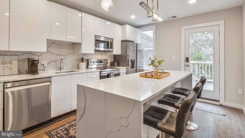 A modern kitchen features sleek white cabinetry and high-end stainless steel appliances, including a refrigerator, stove, and dishwasher. A spacious island with seating is complemented by decorative elements, and natural light streams in through a nearby door leading to an outdoor space.