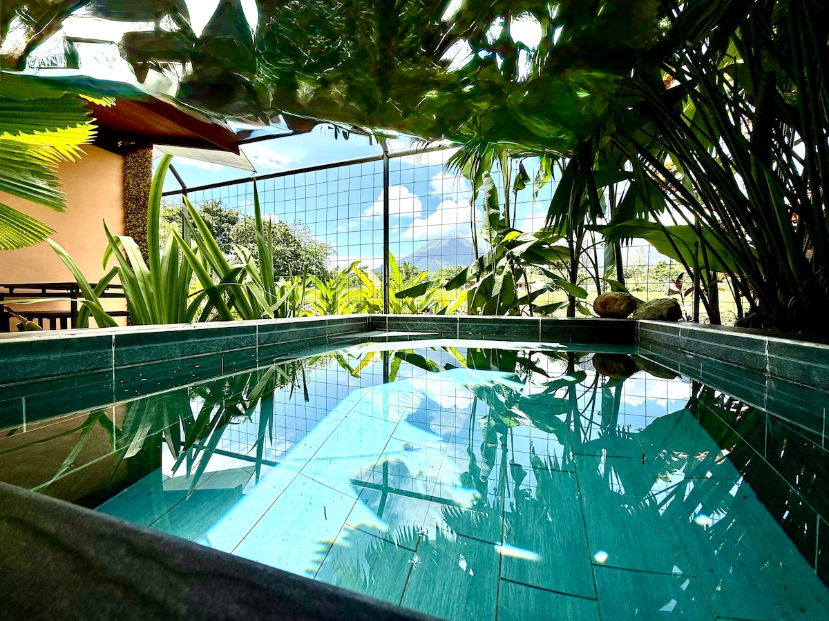A private pool is surrounded by lush green foliage, with reflections of the sky visible on the water's surface. The setting offers a sense of tranquility, complemented by distant mountain views beyond the pool's edge.