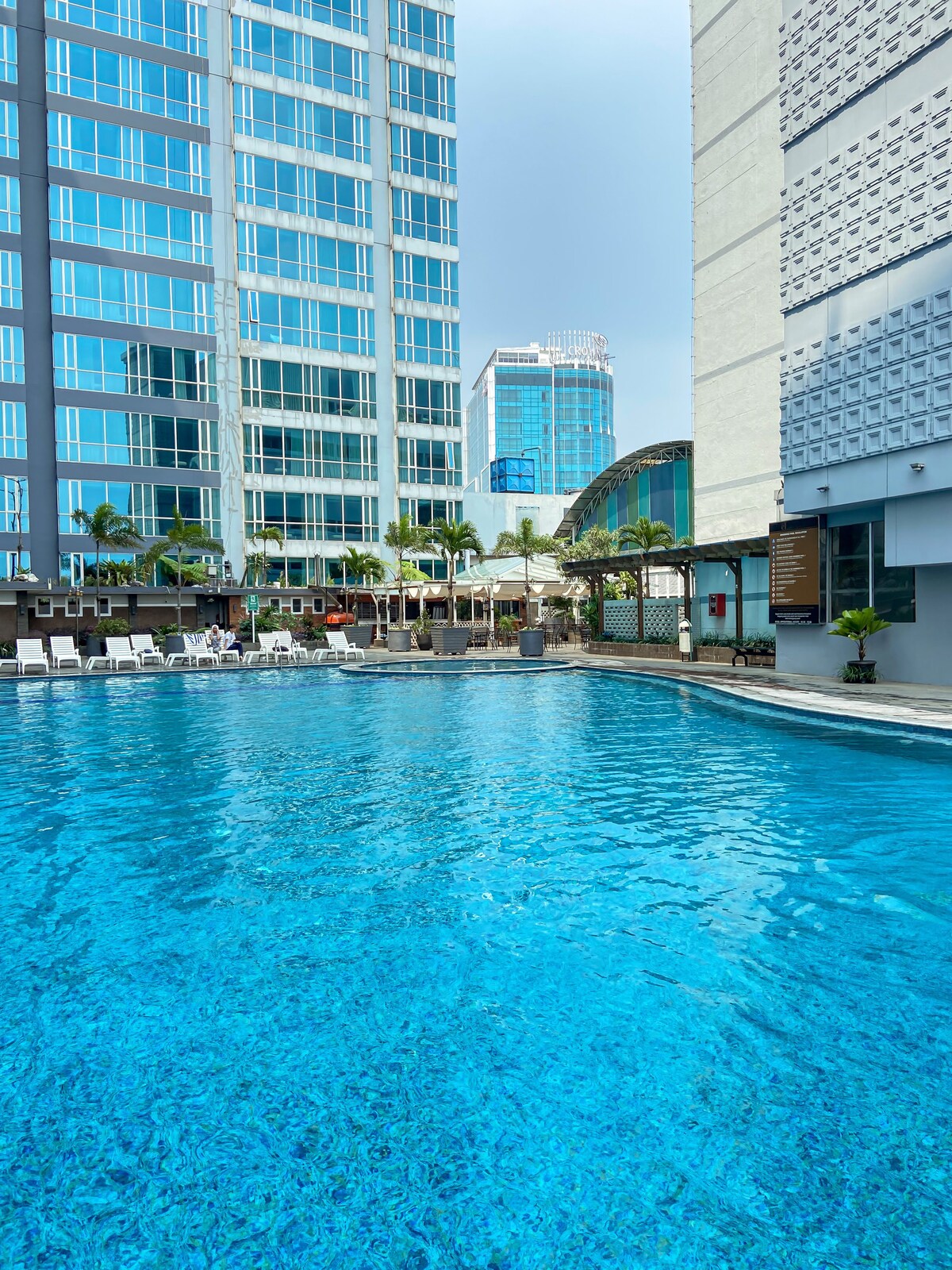A large outdoor swimming pool is surrounded by lounge chairs and palm trees, set against sleek buildings. The clear blue water reflects the vibrant surroundings, creating a serene atmosphere for relaxation.