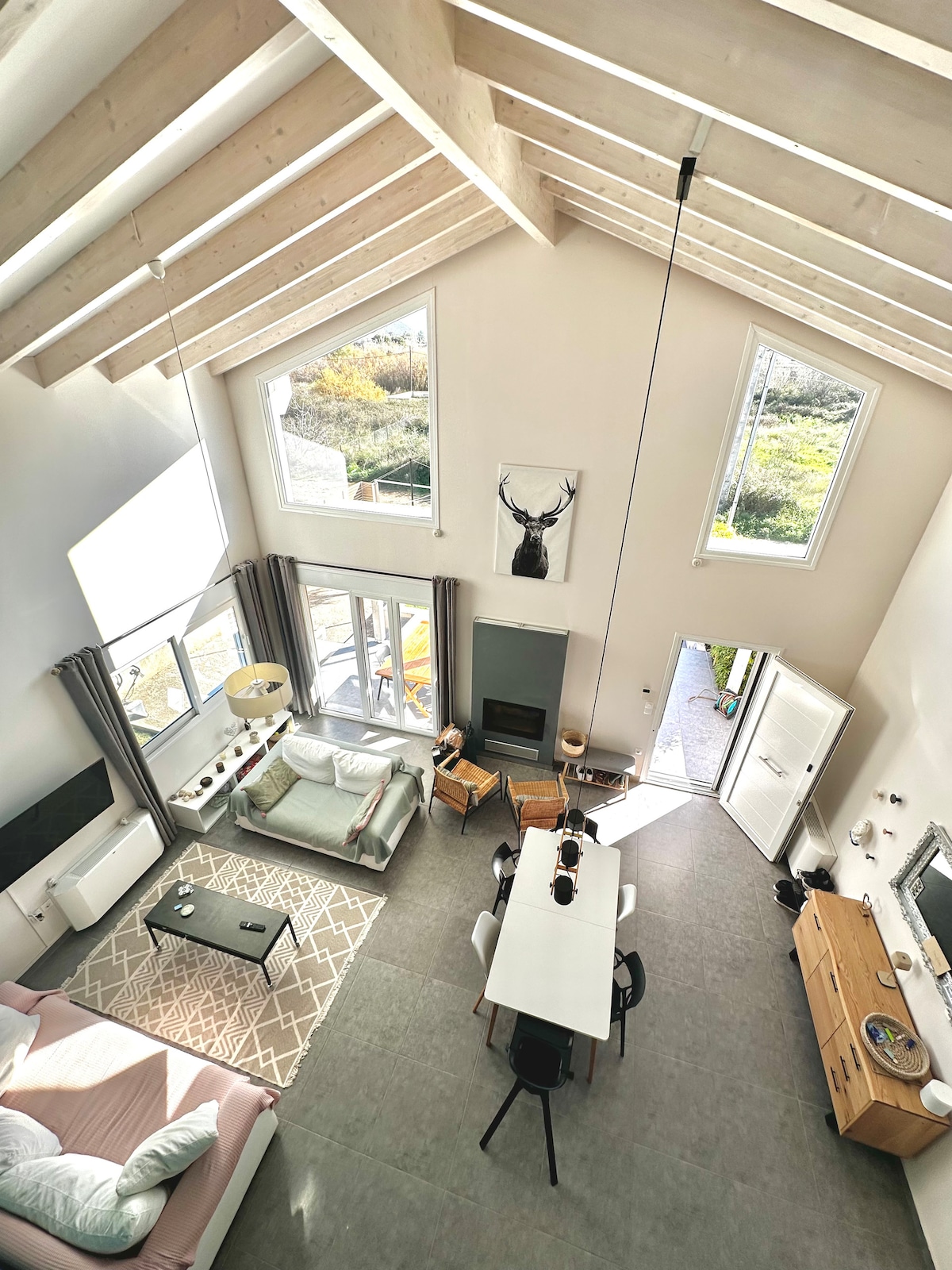 An airy living and dining area is shown from above, featuring high ceilings with exposed beams and large windows that provide natural light. A spacious table is situated near the entrance, surrounded by comfortable seating, while a cozy seating area is visible in the corner.