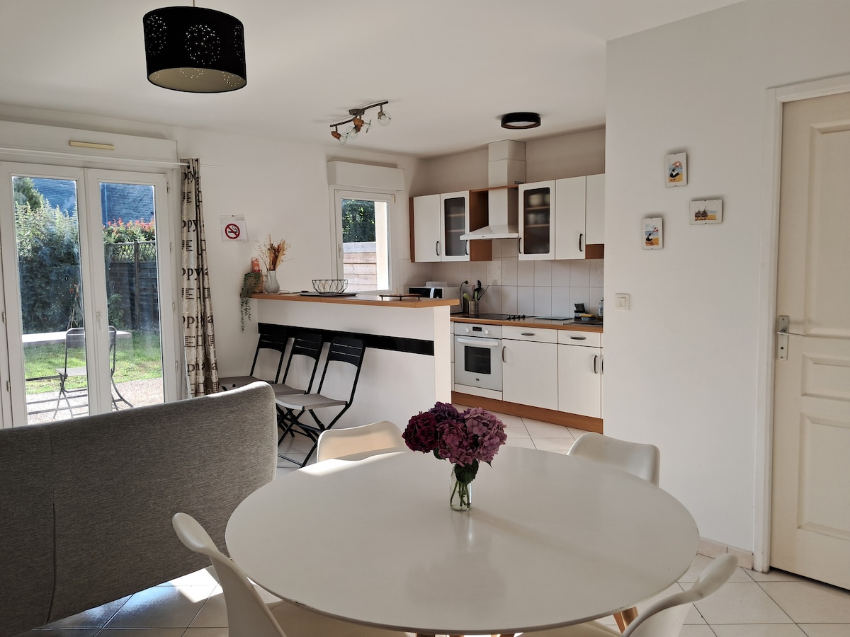 A bright, open living space features a round dining table with white chairs, complemented by a comfortable grey sofa. The kitchen area includes modern cabinetry and appliances. Natural light enters through large glass doors leading to a private, enclosed lawn.