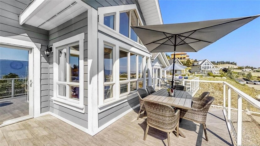 A spacious outdoor dining area is set on a wooden deck, featuring a rectangular table surrounded by comfortable wicker chairs. A large umbrella provides shade, with expansive windows visible in the background, showcasing views of the surrounding landscape.