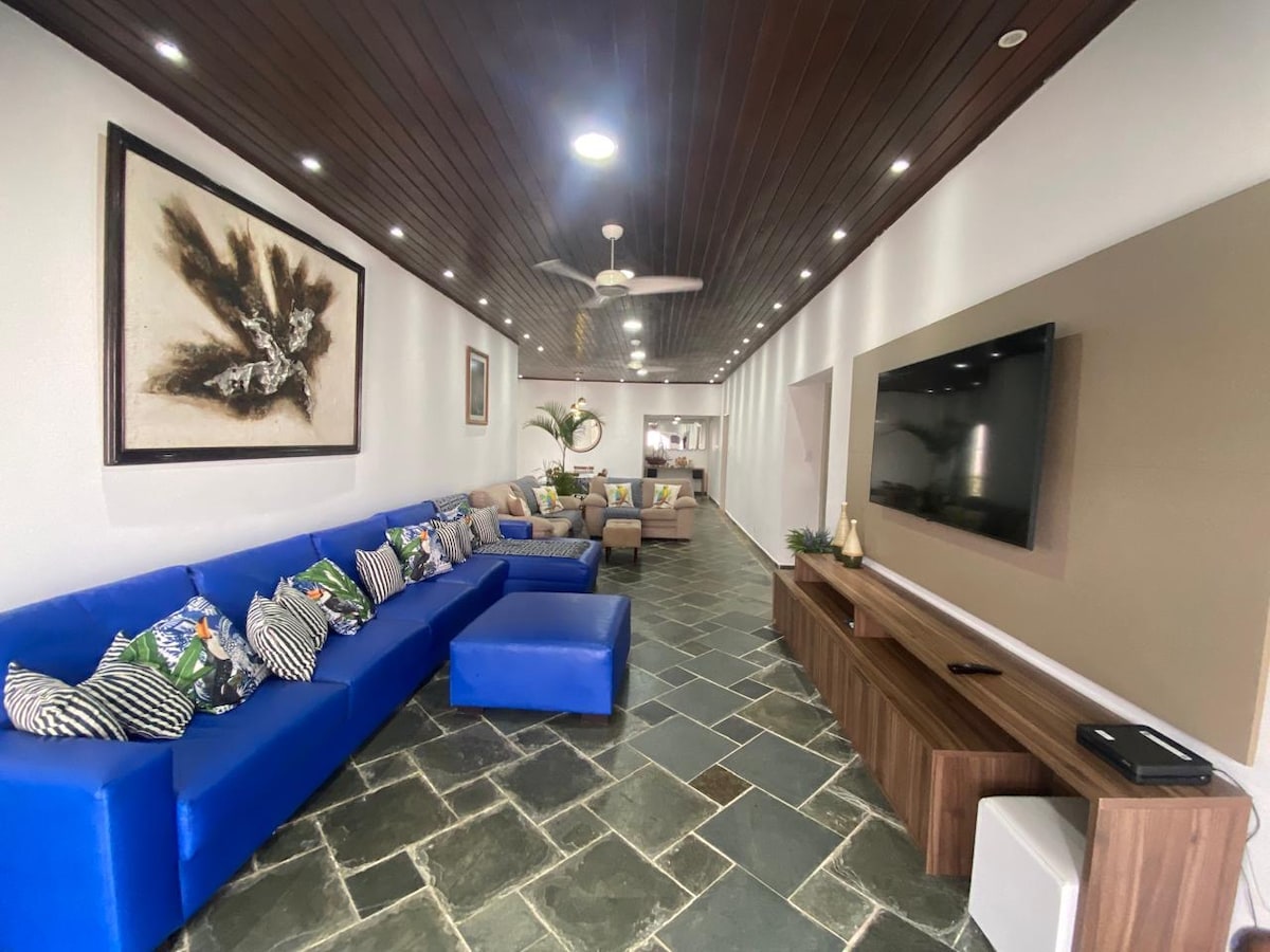 A spacious living area features a large blue sectional sofa adorned with decorative pillows. Light-colored furniture complements the stone flooring. A mounted TV is positioned on a wooden media unit, and energy-efficient lighting is distributed evenly across the ceiling, enhancing visibility.