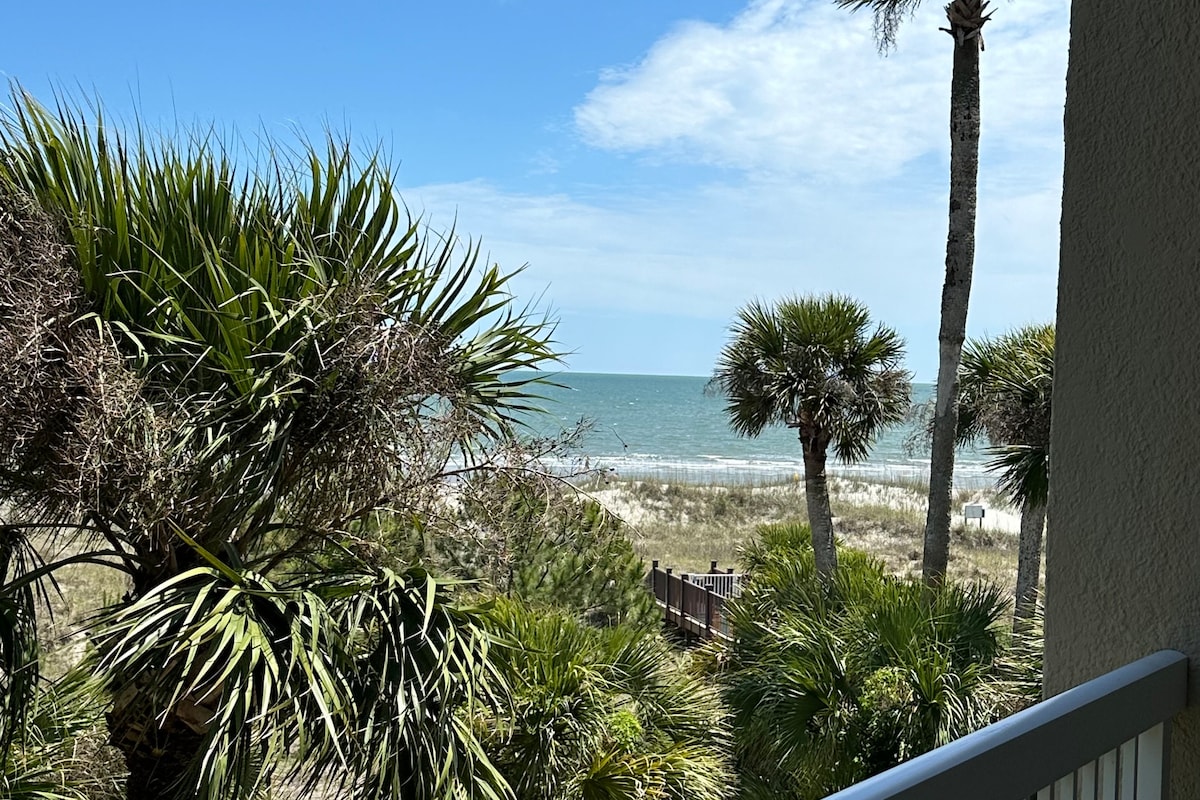 A view from a private balcony showcases lush greenery, including palm trees, against a backdrop of the ocean. The tranquil sea is visible in the distance, meeting the sandy beach that extends along the shoreline under a clear blue sky.