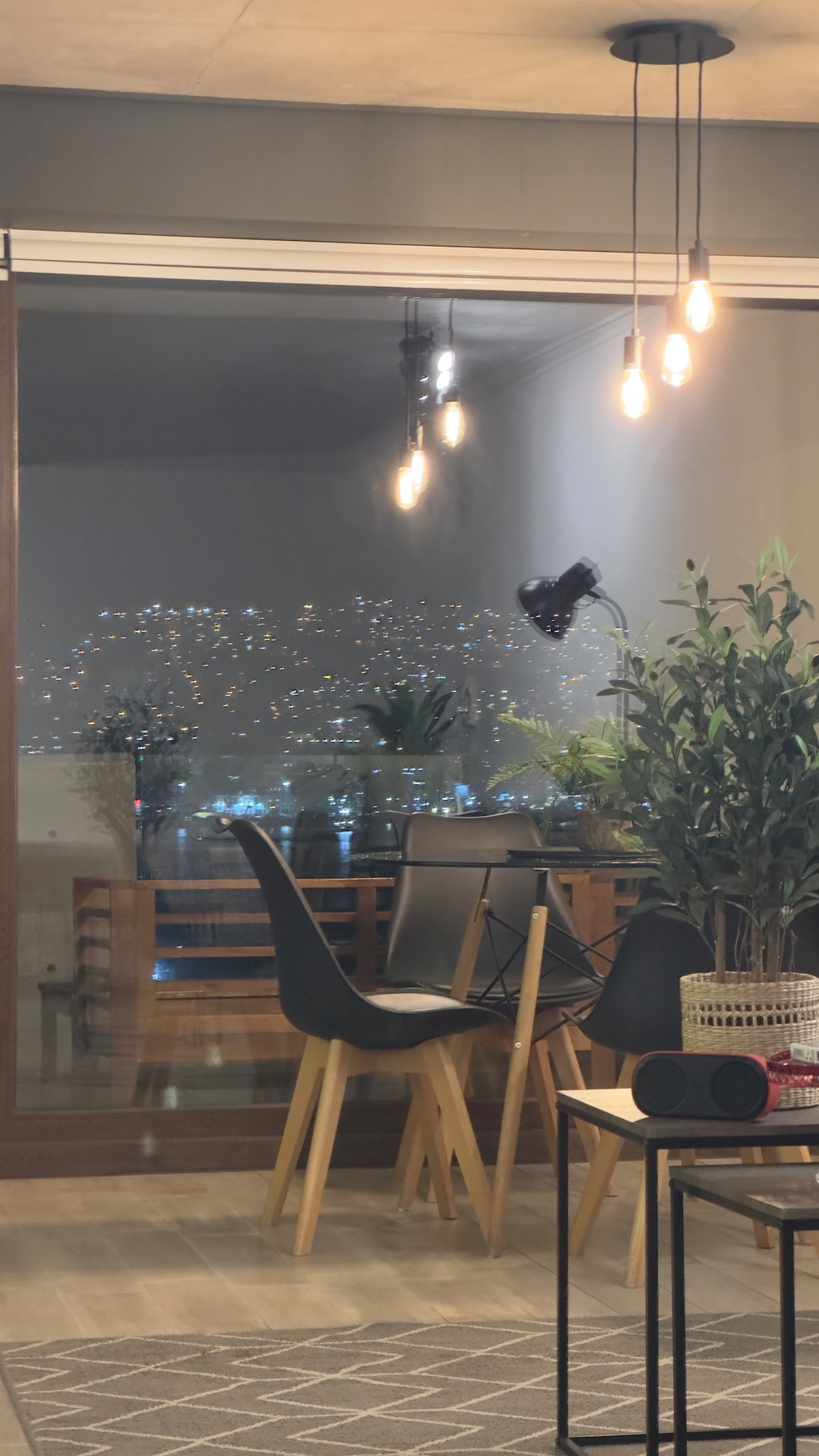 A dining area is shown with modern black chairs and a table overlooking a vibrant cityscape. Warm pendant lights hang from the ceiling, providing a cozy glow. Potted plants are placed near the window, enhancing the serene atmosphere while city lights twinkle in the background.