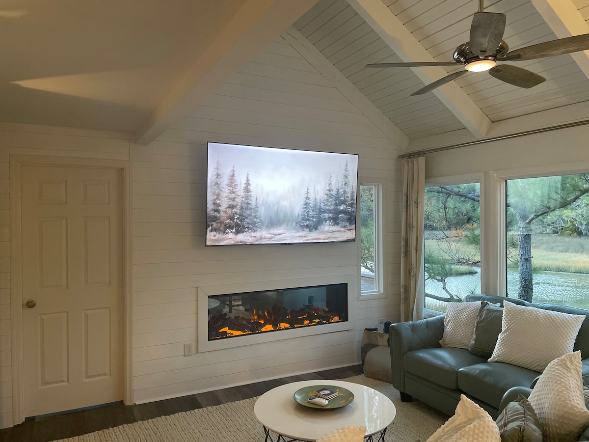 A spacious living area is highlighted by a modern electric fireplace beneath a large mounted television. Windows offer views of the surrounding greenery, while subtle decor creates a relaxing ambiance. A comfortable sofa is positioned to invite seating and leisurely conversations.