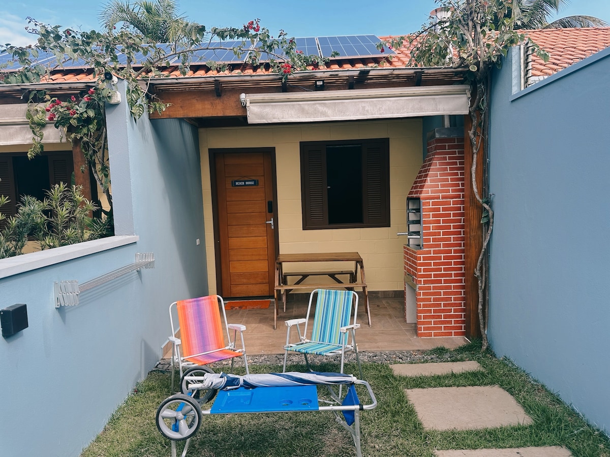 A private entrance is visible, featuring a wooden door framed by a pale blue wall and a charming red-brick barbecue grill. Two colorful lounge chairs are positioned on a grassy area, providing a relaxed outdoor seating option. A bench is situated nearby, promoting an inviting atmosphere.