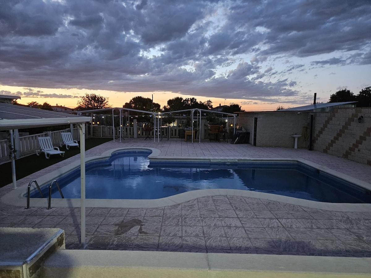 A large swimming pool is surrounded by a tiled patio, with lounge chairs positioned for relaxation. The scene is set against a colorful sunset, revealing a sky with wispy clouds. Outdoor furniture is visible under a covered area in the background.