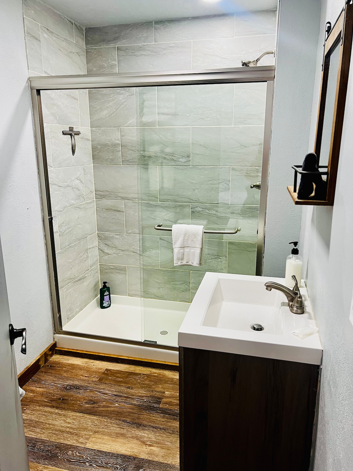 The modern bathroom features a glass-enclosed shower equipped with a sleek chrome fixture. A compact vanity with a smooth countertop is positioned alongside, complemented by natural wood accents. Soft lighting illuminates the space, highlighting the neutral-toned tiles and warm wooden flooring.