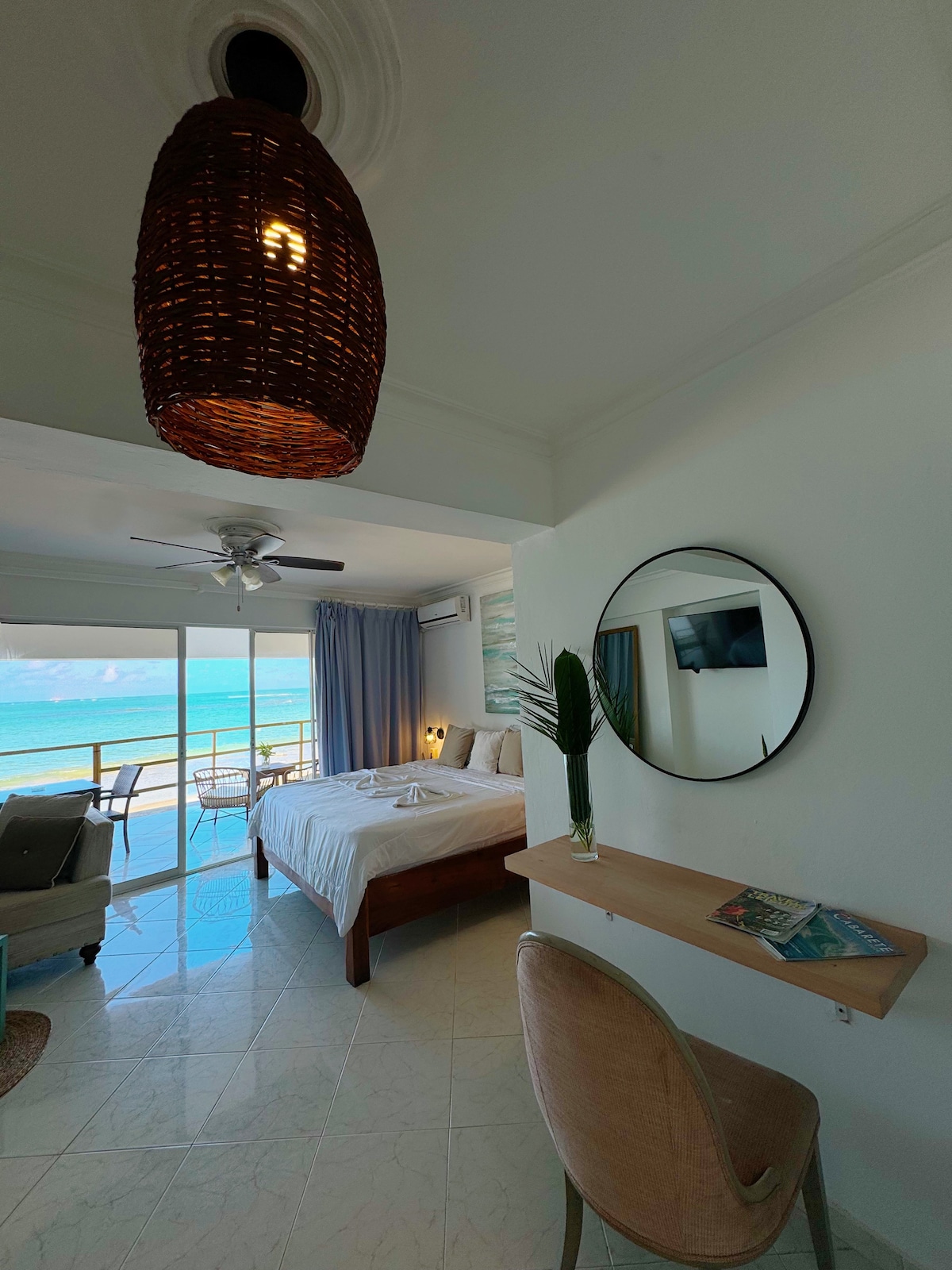 A bright and airy room features a spacious bed, a round mirror above a wooden shelf, and a comfortable chair next to a floor-to-ceiling window. Ocean views are visible beyond the sliding glass doors, while a stylish pendant light adds a decorative touch.