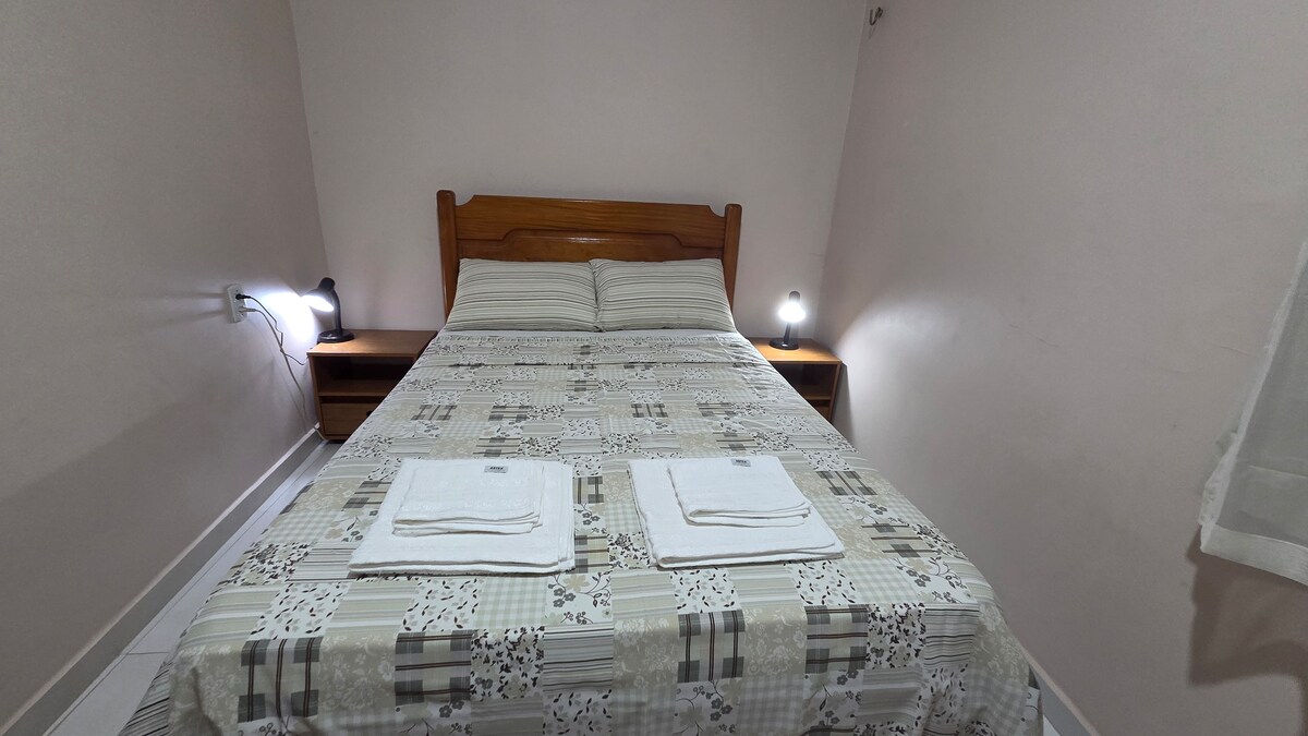 A cozy bedroom is presented, featuring a spacious bed covered with a patterned bedspread. Two bedside lamps provide soft lighting, while neatly arranged towels are placed at the foot of the bed. Soft neutral walls enhance the room's calm ambiance.