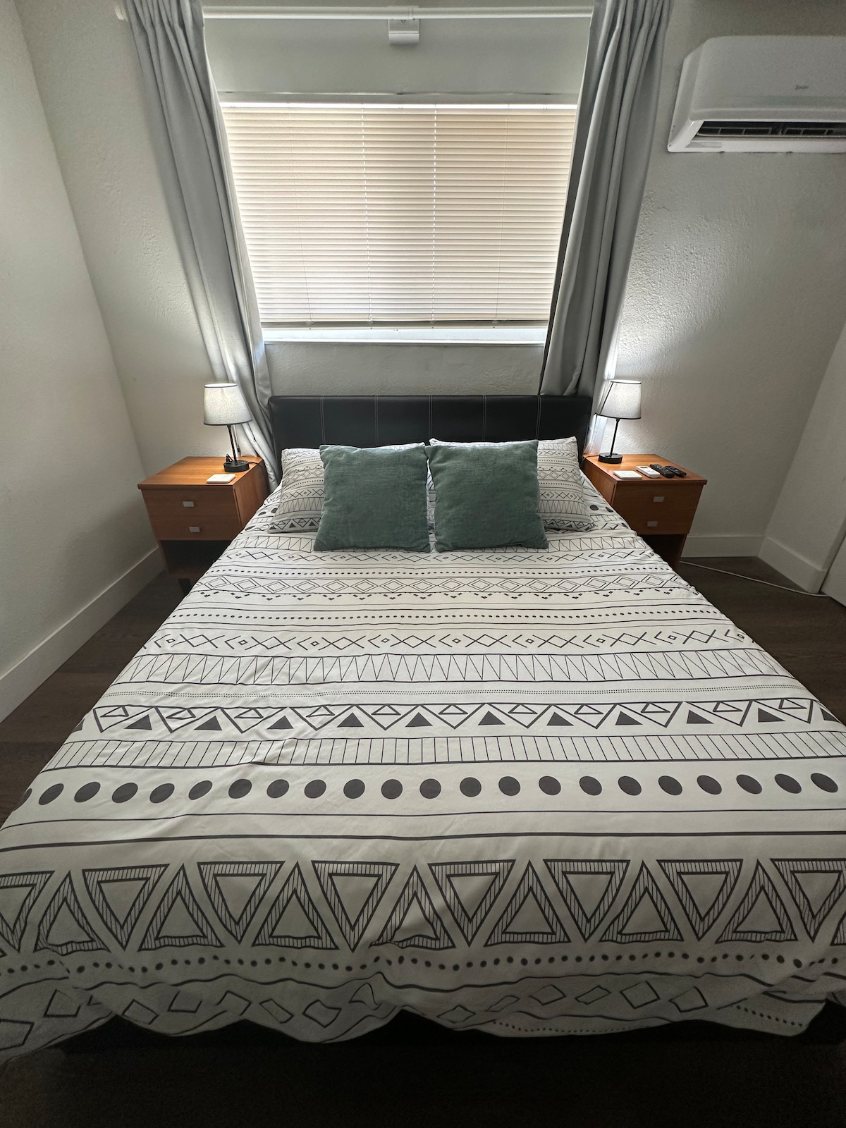 A comfortable sleeping area is centered by a spacious bed, dressed in a patterned comforter. Two bedside lamps provide soft illumination on either side, complemented by small nightstands. Natural light streams through the partially covered window above, enhancing the cozy ambiance.