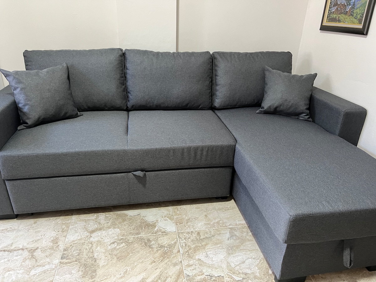 A gray sectional sofa occupies the living area, featuring a comfortable design with plush cushions. Two accent pillows are placed on one side, while the other side extends into a chaise lounge. The neutral tones complement the surrounding light-colored walls.