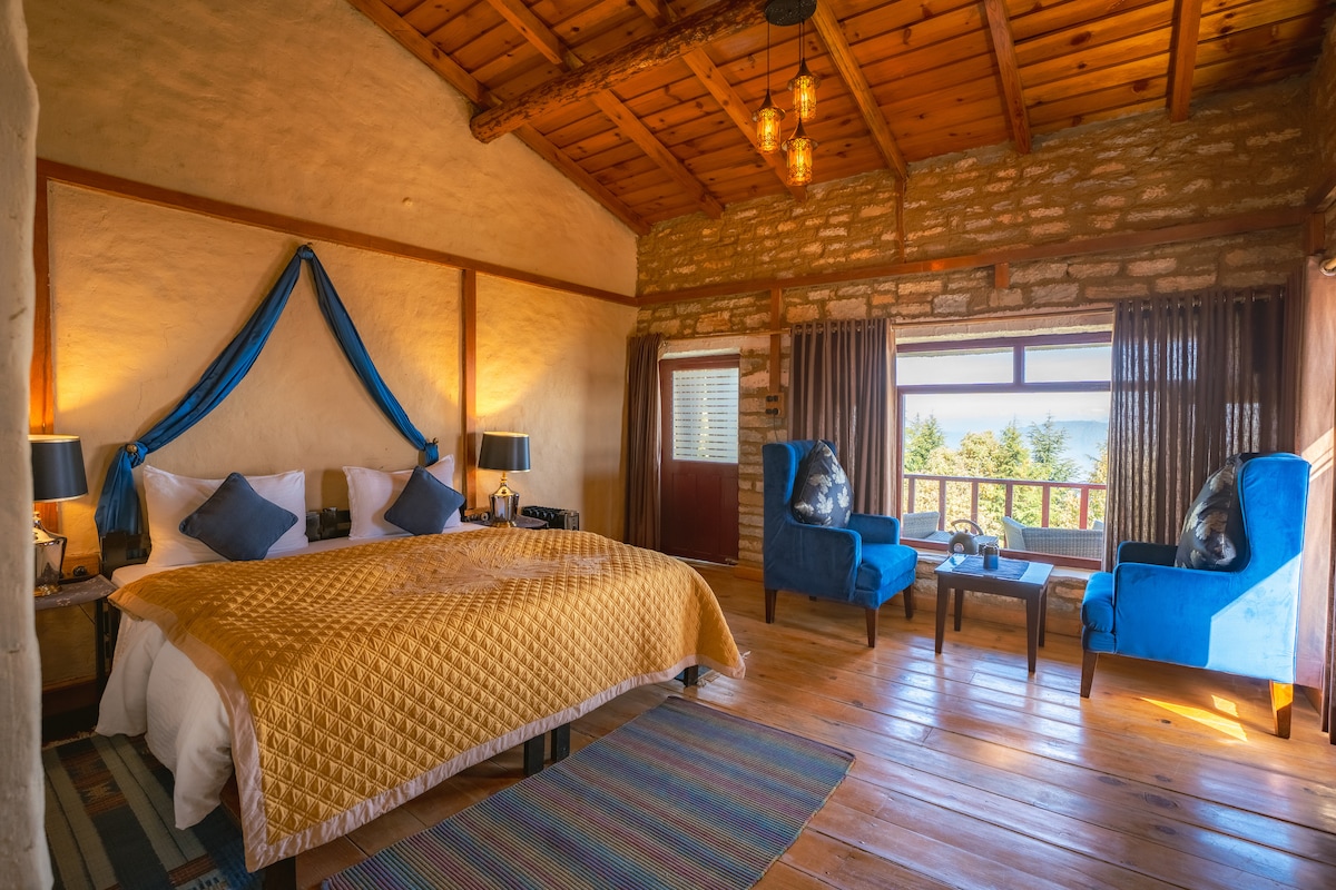 A spacious room features a large golden bedspread over a spacious bed, complemented by blue armchairs and warm wooden flooring. Natural light streams through large windows, offering scenic views of the surrounding forested landscape.