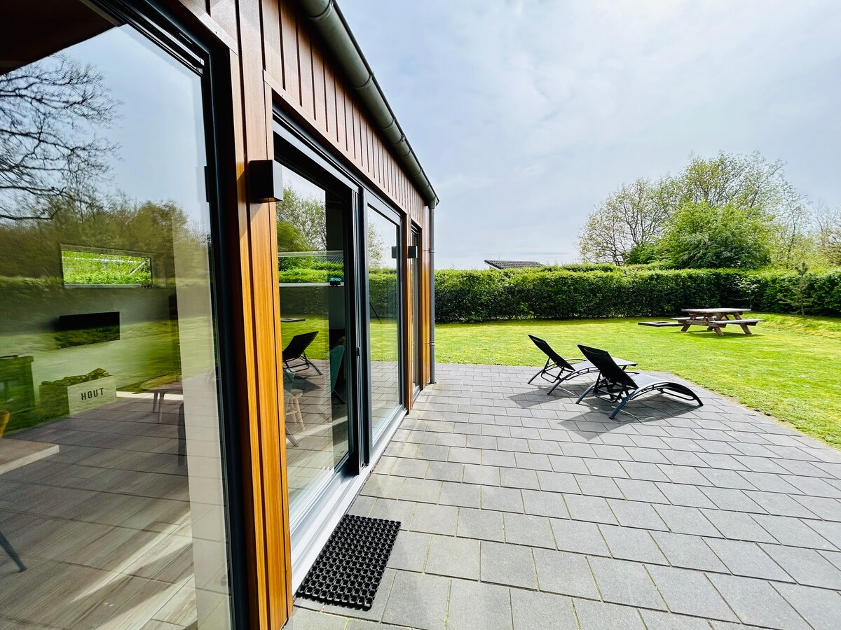 The exterior features large glass doors leading to a spacious patio area. Sun loungers are arranged on a tiled surface, overlooking a grassy yard. A circular picnic table is positioned in the yard, surrounded by hedges and trees, providing a serene outdoor space.