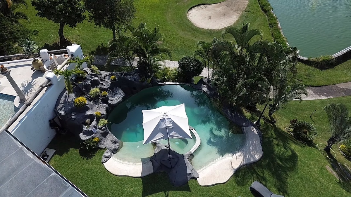 A solar-heated pool features a sandy entry and a nearby bubbling jacuzzi, surrounded by lush greenery. A large umbrella provides shade over lounging chairs. The pool area offers views of a golf course and a tranquil lake, enhancing the outdoor relaxation experience.