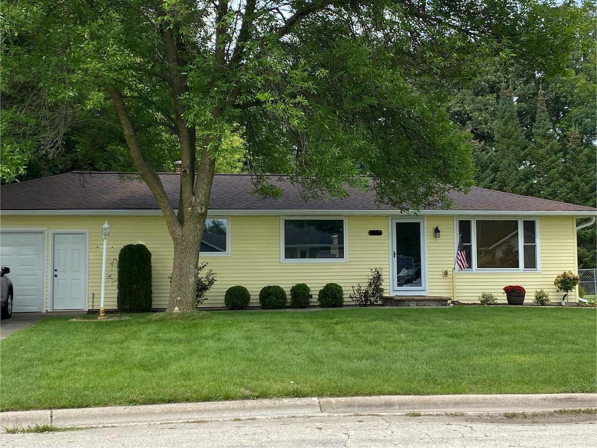 A single-story home is displayed with a yellow exterior and a dark roof. The front yard features well-manicured grass and bushes, while an American flag is visible near the entrance. A garage is present on the left side of the house.