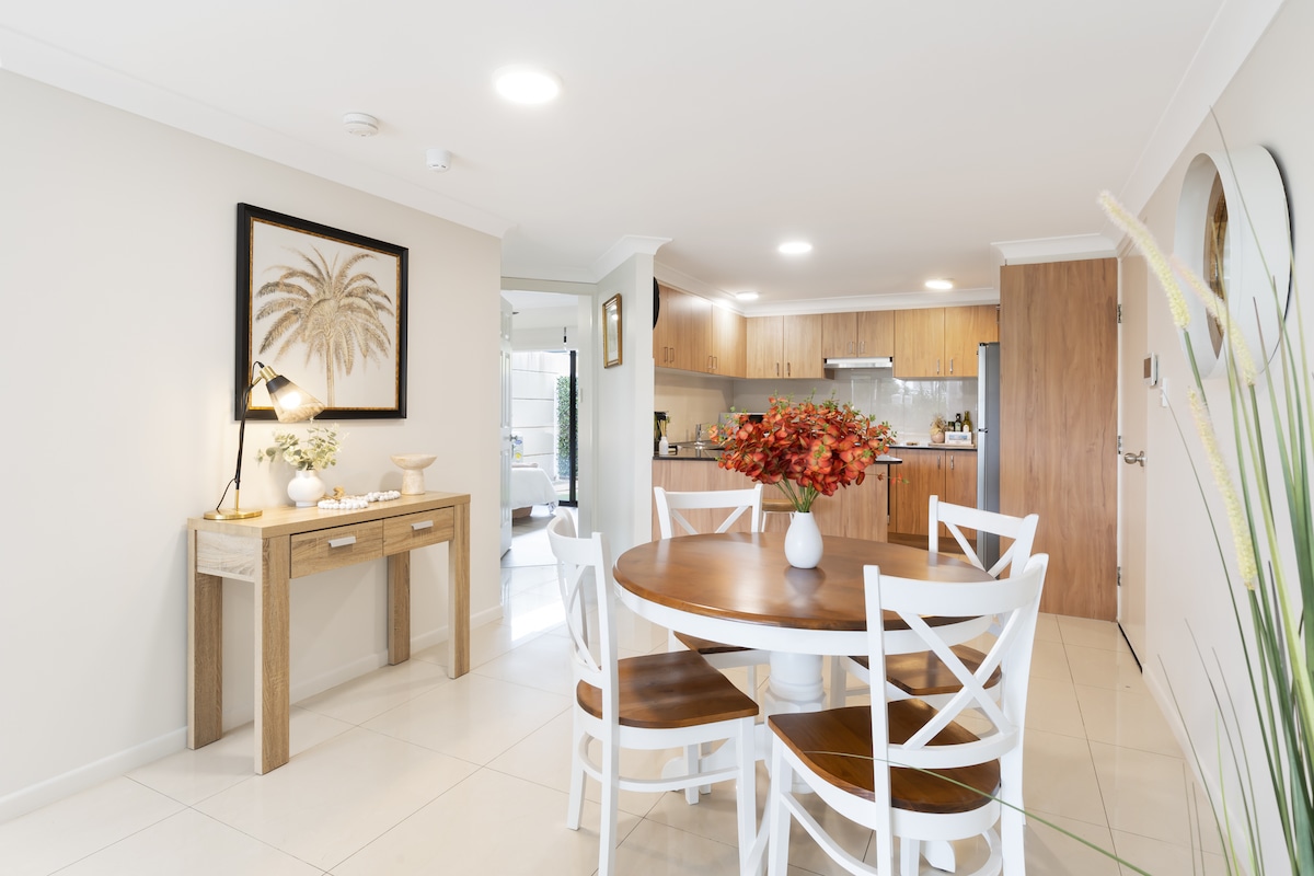 A modern dining area features a round wooden table surrounded by white chairs. A console table with decor stands against a wall adorned with a framed palm print. The open layout connects to a kitchen area, illuminated by soft ceiling lights.