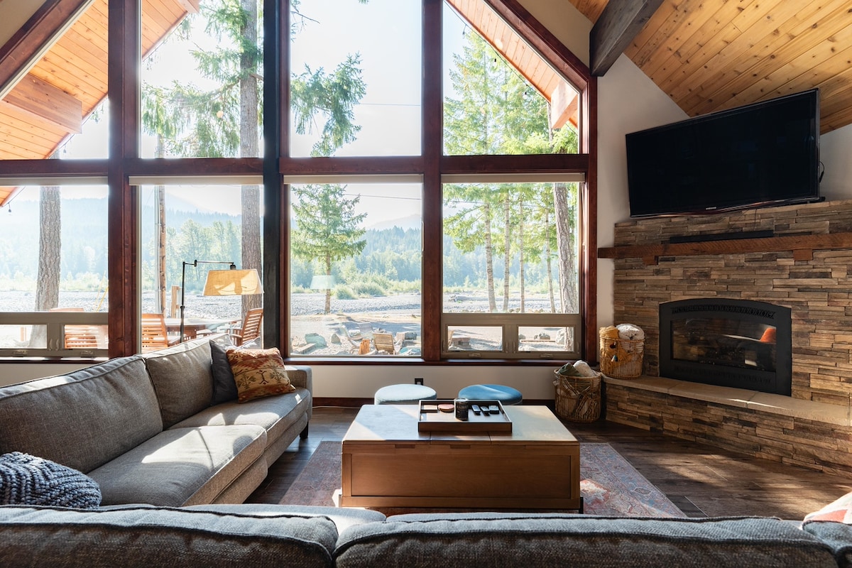 The spacious living area features large windows that offer views of the surrounding valley and river. A comfortable sectional sofa and a wooden coffee table are arranged to create a welcoming space. A gas fireplace is positioned against a stone wall, adding warmth to the area.