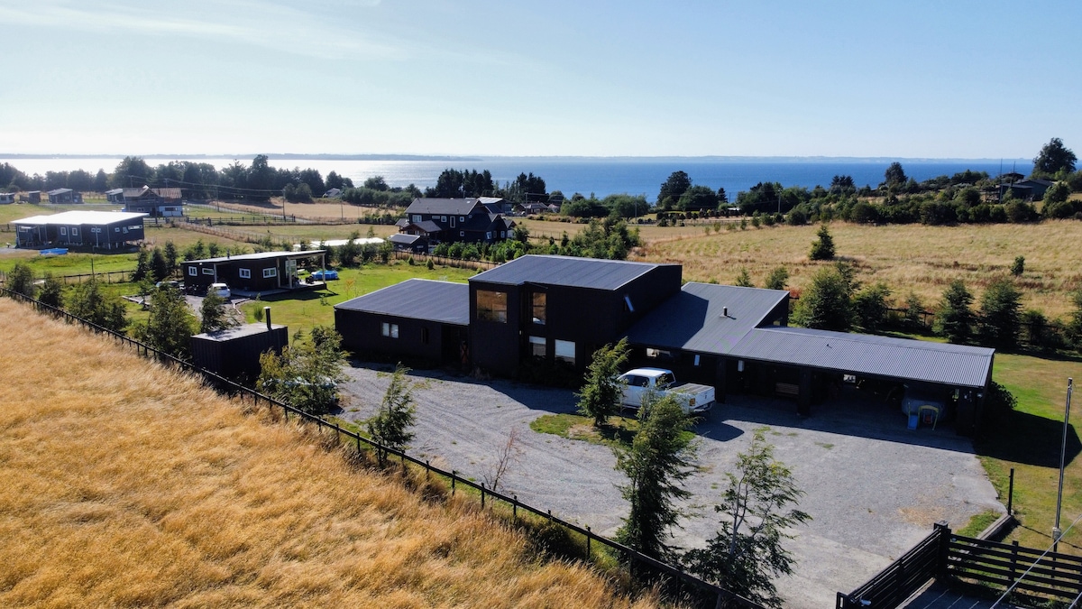 An aerial view presents a modern dark-wood house surrounded by lush green fields and a gravel driveway. The coastline is visible in the background, with the expansive blue water complementing the serene setting. Trees and various vehicles add to the rural charm.
