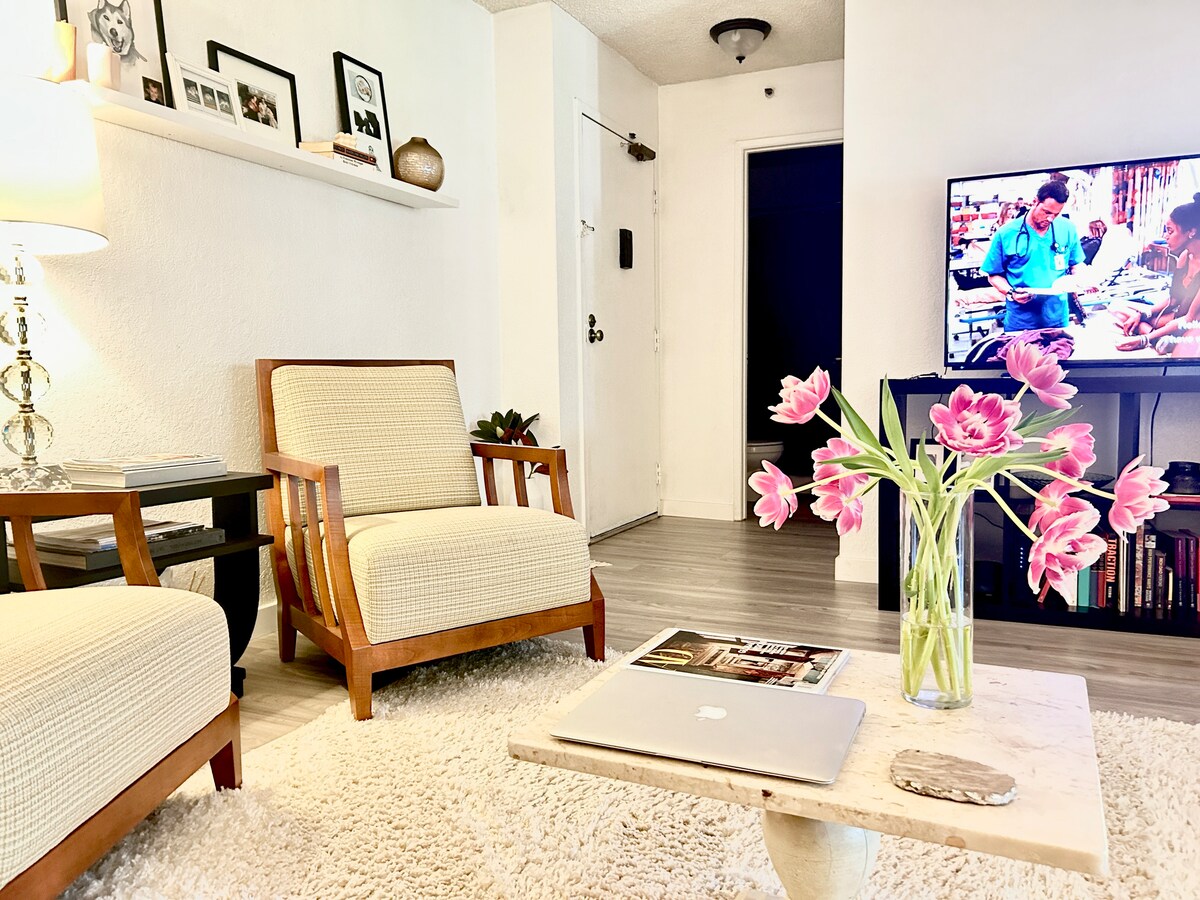 A comfortable living area features two beige armchairs positioned near a coffee table with a laptop and magazines. A vase of pink tulips adds a touch of color. A large screen TV displays a scene, while the entrance door and bookshelf are visible in the background.