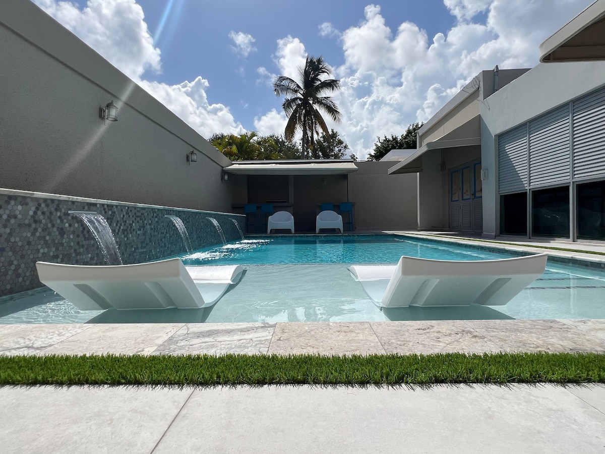 A modern salt-water pool features two stylish loungers partially submerged in shallow water. Water cascades from a decorative wall, surrounded by a clean stone deck. Tropical landscaping is visible in the background, with palm trees under a bright sky dotted with clouds.
