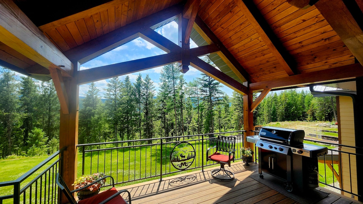An outdoor patio area features a black propane BBQ and two red chairs under a wooden pergola. Surrounded by trees, the space offers an expansive view of the well-maintained lawn and forest beyond, inviting relaxation and outdoor dining.