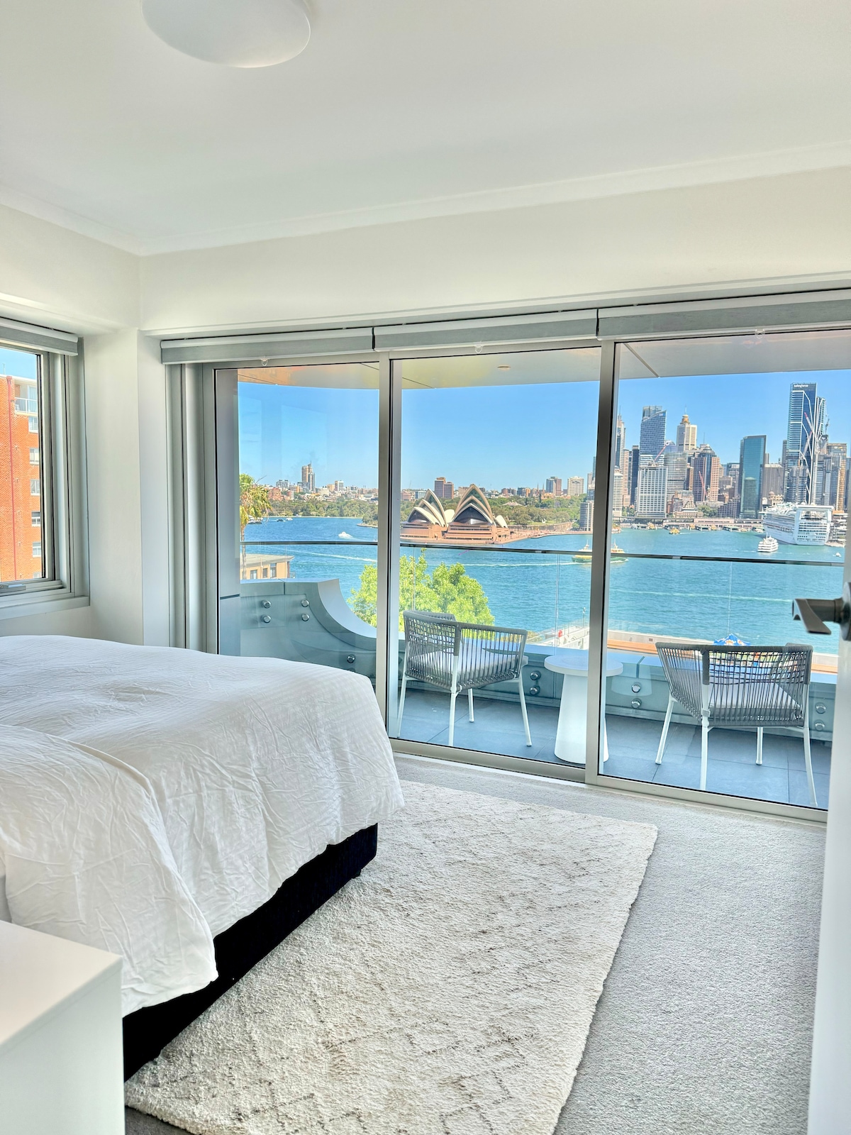 A bright bedroom is highlighted by large glass sliding doors that showcase a stunning view of the Sydney Opera House and city skyline. A spacious bed is set against a soft area rug, with natural light flooding the room from both windows.
