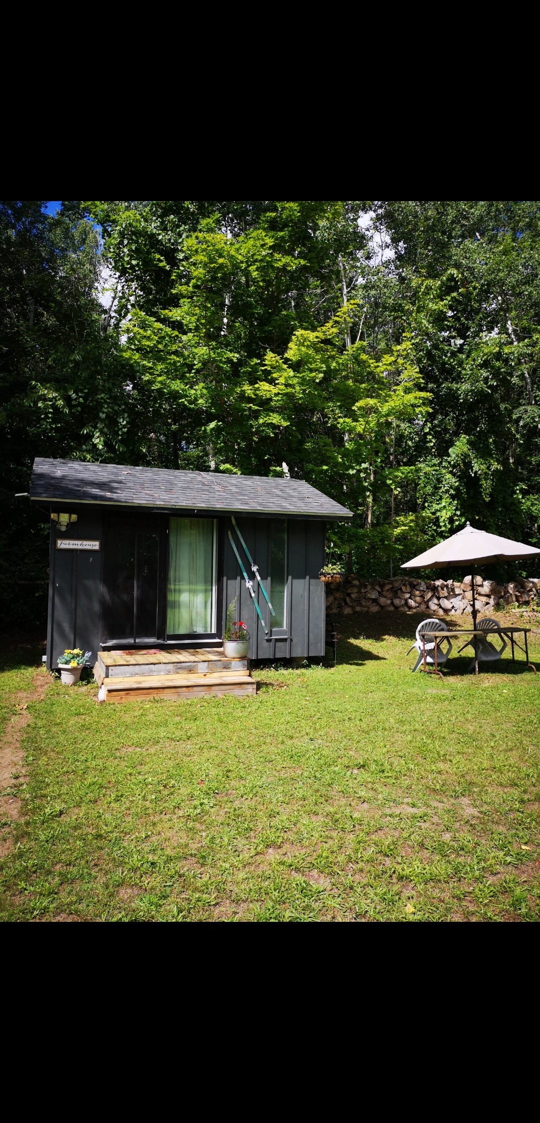 A cozy glamping cabin with a dark exterior is set in a grassy area surrounded by tall trees. A small wooden deck leads to the entrance, while a circular table with an umbrella is positioned on the side, offering a shaded outdoor seating area.