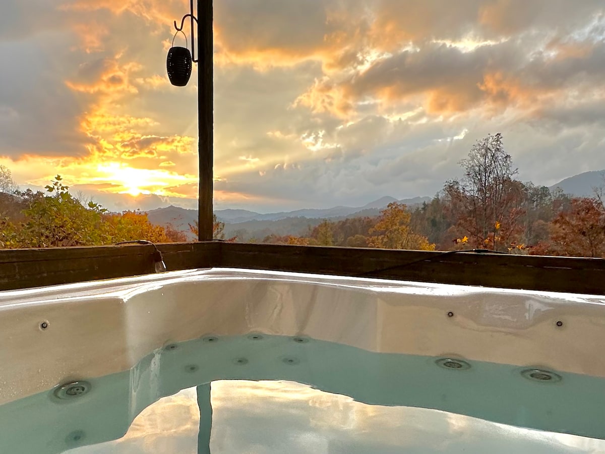 A hot tub is positioned on a covered balcony, offering views of rolling hills and a vibrant sunset. The tranquil water reflects hues of orange and gold, while the surrounding landscape features a mix of trees in autumn colors.