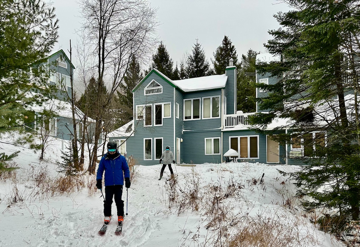A person skis down a snowy path in front of a two-story blue condo surrounded by evergreen trees. The building features large windows and a balcony. A second skier is visible in the background, with snow-covered terrain enhancing the wintry scene.