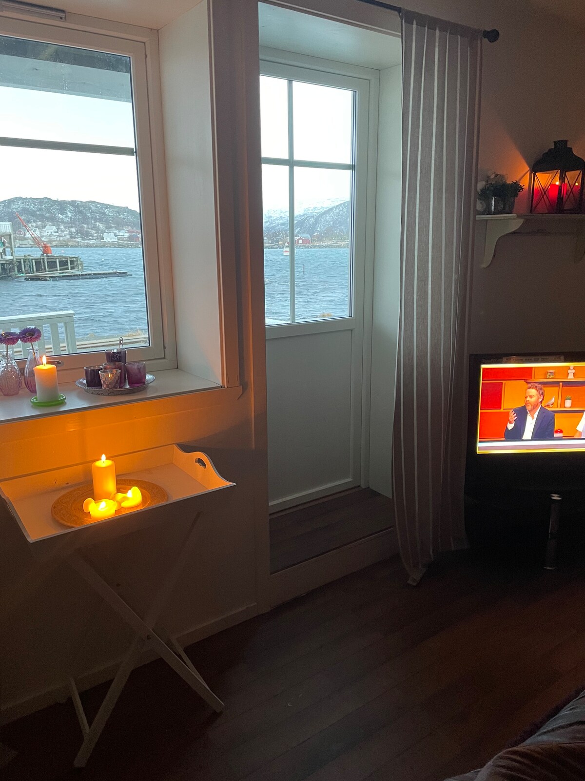 A cozy corner of the living area features a window with a view of the water and surrounding hills. A small tray holds lit candles, while a television displays a news program. Sheer curtains allow natural light to filter in.