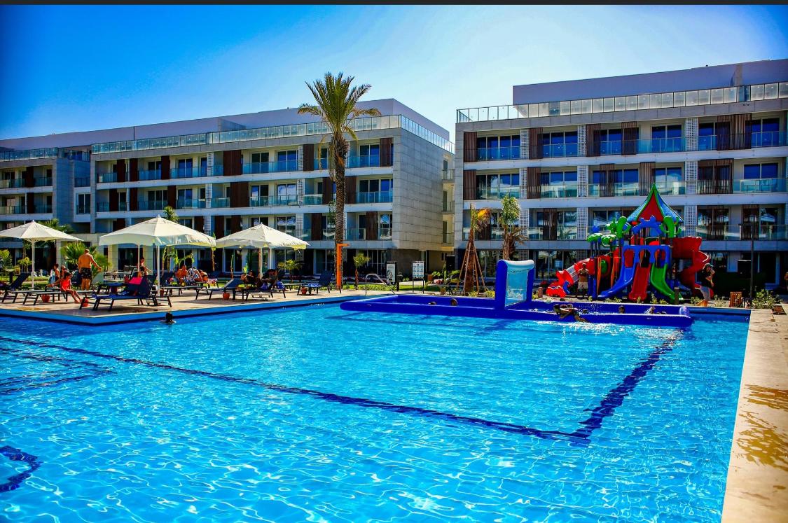 A large swimming pool is surrounded by lounge chairs and umbrellas. In the background, multiple balconies overlook the pool area. A colorful children's play structure is positioned nearby, adding a playful element to the landscape. Lush palm trees provide a tropical ambiance.