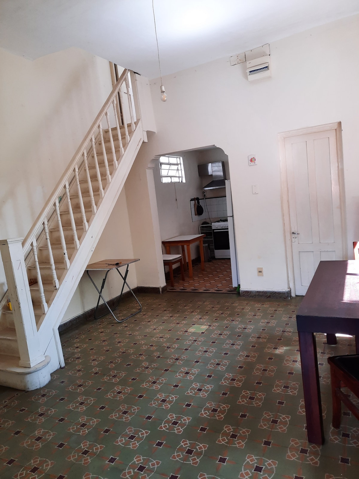 A spacious and airy living area features tiled flooring with a geometric pattern, complemented by a wooden table and chairs. A staircase ascends to an upper level, while a glimpse of the kitchen is visible through an archway. Natural light enters through a window.