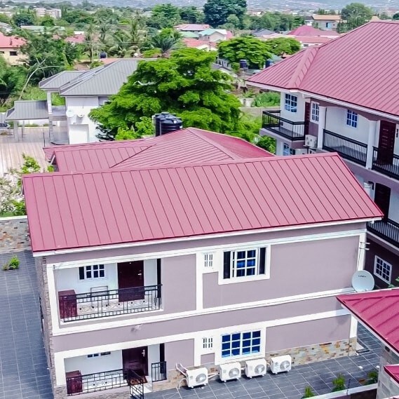 The image showcases an aerial view of a charming apartment building with a red roof. The structure features multiple balconies and white accents. Surrounding greenery is visible, enhancing the tranquil atmosphere of the neighborhood.
