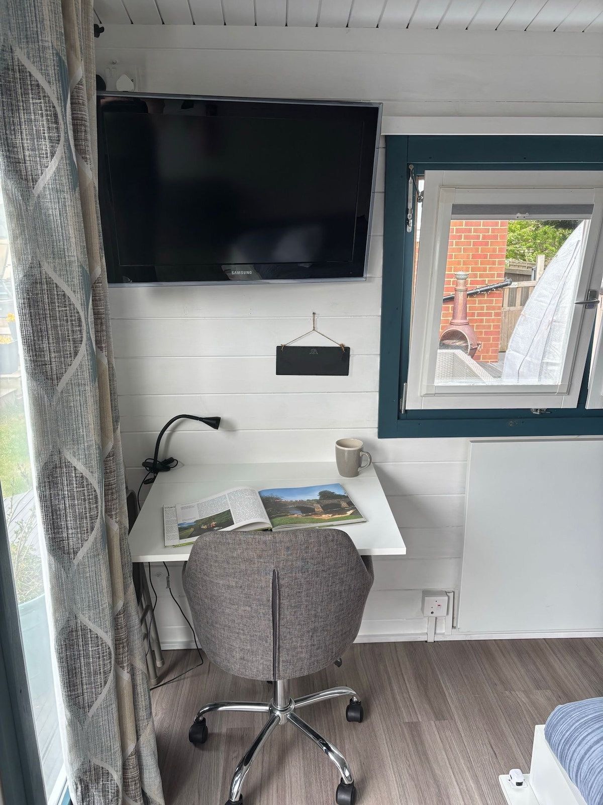 A work and dining area is featured, showcasing a compact table with a magazine and mug. A comfortable, grey chair is positioned at the table. A flat-screen TV is mounted on the wall, and a window allows natural light to fill the space.